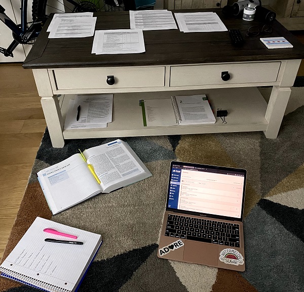 A low coffee table scattered with stacks of white papers surrounded by a school notebook, text book and open laptop